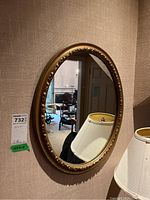 Oval mirror with ornate gold composite frame hanging on wall, reflecting a room interior with furniture and lamps.