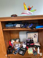 Office supplies and electronic items arranged on wooden shelves, including adding machine, stapler, hole punch, eyeglasses, cases, CD player with headphones, and birthday hat.
