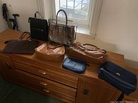 Seven purses and backpack arranged on a wooden dresser, showing various colors and styles including metallic brown croc-embossed purse, black backpack, designer purses