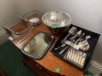 Photo of silver plate trays, bowls, and basket on wooden table showcasing the reflective surfaces and overall condition.