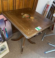 Square wooden table with floral design on top, beveled edges, and pedestal legs shown from an angled side view.