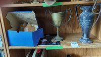 Shelf showing two glass food covers in a blue box and two trophies on a wooden shelf.