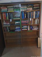 Pair of two modern wooden bookshelves filled with various books from front view under ambient indoor lighting.