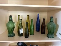 Nine coloured glass bottles displayed on shelf including two large eagle-shaped green glass bottles and seven traditional shaped bottles in various colours and sizes