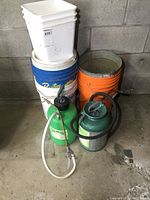 Two garden sprayers in front of stacked plastic buckets in blue, white, and orange.