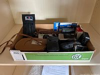 Overview of cameras and equipment in cardboard tray showing Brownie movie camera, Konica cameras, bags, manuals, and chargers.