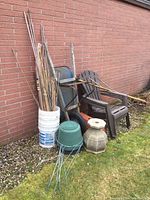 Photo showing all items against brick wall: two resin Muskoka chairs, older wheelbarrow, various garden stakes in buckets, plastic planters, and concrete planter on grass.