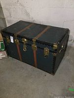 Front and top view of large black metal trunk with brown straps and brass corner protectors.