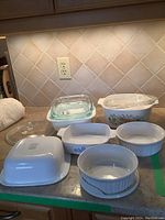 Seven Corningware casseroles and lids on kitchen counter, showing white square, fluted round, Cornflower and Spice of Life patterns.