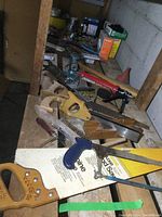 Shelf with assorted staining and wood-handled saws, blue hacksaw, wooden mallet, caulking gun, small vise, and other hand tools laid on a wooden surface