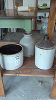 Three ceramic vessels displayed on a wooden shelf including a crock jug, a round pot, and a jug with a dark rim.