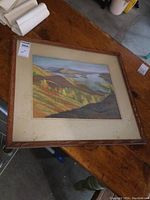 Framed signed original landscape art lying on wooden table showing whole framed piece and mat with glass covering.