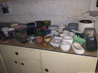Wide view of assorted small planters, pottery, glass, and plastic ware on a cabinet showing variety of shapes, sizes and colors