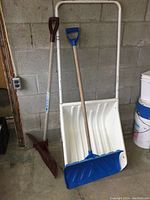 Three snow shovels shown standing against a concrete wall: one large white plastic snow scoop with metal handle, one red plastic push shovel, one blue plastic push shovel.