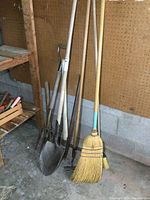 Photo showing shovel, fork, hoe, sledgehammer, pickaxe, and two brooms leaning against a pegboard wall in a storage area.