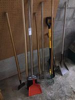 Full view of yard tools arranged against wall, showing shovels, rake, edger, hoe, broom, and dustpan.