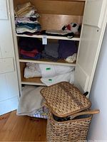 Photo of wooden shelving with folded bedding items and plush panda hot water bottle cover, above woven laundry hamper and blanket on floor