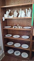 Wooden shelf with nine white ceramic nativity plates arranged on four shelves; small white ceramic nativity figurines on upper shelf