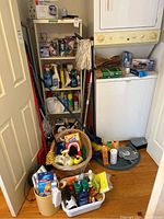 Photo of closet with mops, brooms, cleaning supplies on shelf, wicker basket, small heater, dish pan, ironing board, and waste bin