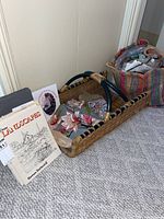 Woven basket with handle containing embroidered fabric and embroidery thread, adjacent to large straw tote filled with wool and threads, along with instructional drawing books and vacuum storage bags.