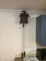 Front view of full cuckoo clock mounted on wall showing carved wooden housing, clock face, two metal weights, and chains.