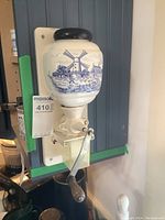 Front angle view of coffee grinder mounted on wall showing ceramic container with blue Delft windmill design, metal crank handle with wooden knob, and white backing plate.
