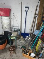 Wide shot of various garden tools including shovels, concrete temple, planters, white plastic pails, wire basket, green portable garden seat, and seed starter trays.