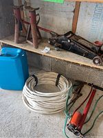Two red metal bottle jacks, black hydraulic floor jack, blue plastic jug, coiled white wire, orange foot pump, and red battery cables on a wooden shelf and floor.