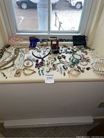 Wide view of assorted jewellery arranged on a window ledge including beaded necklaces, metal chains, bracelets, earrings, and pins.