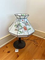 Full view of the lamp showing the stained glass-look shade with Asian figures and nature scenes, placed on a wooden floor near wall corner.