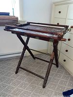 Folding luggage rack shown with woven tray installed, displaying carved wooden legs, brass rods, and visible wear on the woven top.
