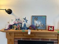 Wooden mantel with framed needlepoint, glass cat figurines, wooden cats, fabric ornament, stone owl and notepad holder displayed