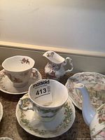 Photo showing three teacups and three saucers with seasonal and wedding-themed illustrations, plus a Wedgwood plate in the background.