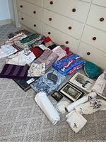 Photo showing a variety of folded tablecloths, runners, placemats and decorative linens in different colors and patterns including reds, blues, and floral motifs spread on carpet.