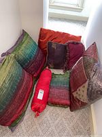 Photo showing all eight throw pillows arranged on a carpeted floor corner, including multicolored patterned pillows, solid orange, purple pillow with fringe and red bolster pillow.