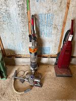 Dyson Slim DC18 upright vacuum and Dyson DC61 handheld vacuum with charging cord in garage setting on concrete floor against insulation wall