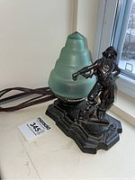 Overall side view of vintage bronze base lamp with man figurine and green frosted glass bulb on a window ledge.