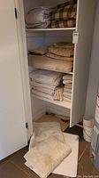 Closet shelves with stacked folded sheets, towels, and blankets in cream and beige colors. White and cream bath mats are on the floor.