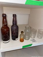 Full lot on white shelf showing two brown bottles, small clear bottle with liquid and HH marked gold lid, a textured Bireleys glass bottle, and two clear beaker glasses.
