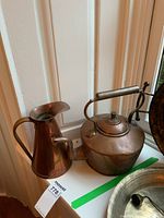 Copper pitcher and kettle displayed together on a windowsill with natural light, showing overall appearance and relation in size.