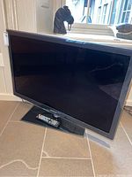 Front view of Samsung TV on stand with remote on the base, showing screen and bezel.