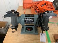 Front view of Delta bench grinder showing the grinding wheels, protective eye shields, and orange power cord on wooden table.