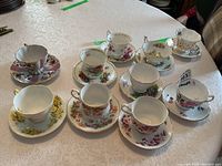 Photo of 8 assorted floral pattern teacups and saucers on a white tablecloth showing variety in pattern and color.