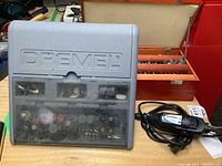 Grey plastic case of assorted Dremel bits and rotary tool lying next to orange metal accessory case.