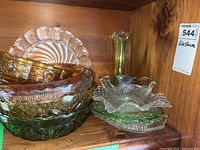 Wooden shelf holding a collection of various glassware items stacked and arranged including an amber bowl, green glass fruit bowl, clear ribbed bowl, and a green slender vase with floral top.