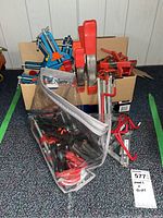 Full view of lot showing cardboard box holding various clamps with red handles, blue corner clamps, and a plastic bag containing assorted smaller grip clips and hooks.
