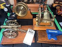 Photo showing black decorative microscope, brass Royal Mariner clock, brass mounted bell on wood, brass mounted cannon on wood, and blue Benchmarked anvil on wood base.