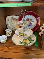 Three tier pastry server with floral design and two decorative plates behind it. Creamer and sugar bowl with yellow flower and gold trim to the left.