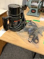Black & Decker router with power cord and black handle, placed on a wood surface with a pair of scissors in a plastic bag beside it.