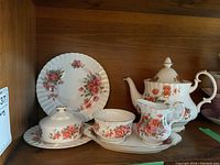 Photo of the serving set pieces including teapot, tube bowl, creamer, butter dish plate and teacup on saucer on a wooden shelf